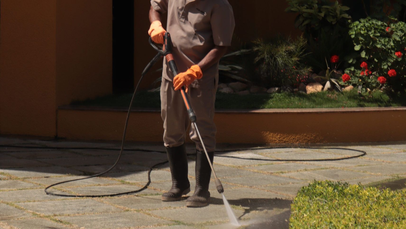 A gardener wearing a hat cleans an urban sidewalk with a pressure washer.